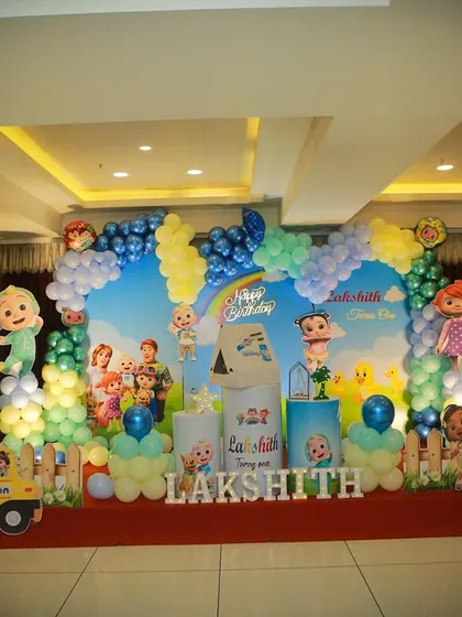 A large-scale Cocomelon theme decoration for a party hall, with a full stage setup including a backdrop, character props, and balloon pillars.