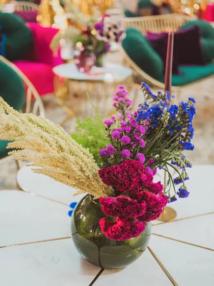 A small, vibrant floral arrangement in a green glass vase, showcasing a mix of textures and colors with pampas grass and fresh blooms.