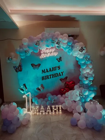 A beautiful butterfly themed first birthday party. The circular backdrop is surrounded by a pastel balloon arch, with a neon 'Happy Birthday' sign, marquee number '1', and light up letters for the name 'Maahi'.