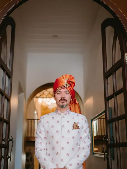A striking portrait of the groom in a white sherwani and a vibrant orange safa, framed perfectly in an arched doorway.