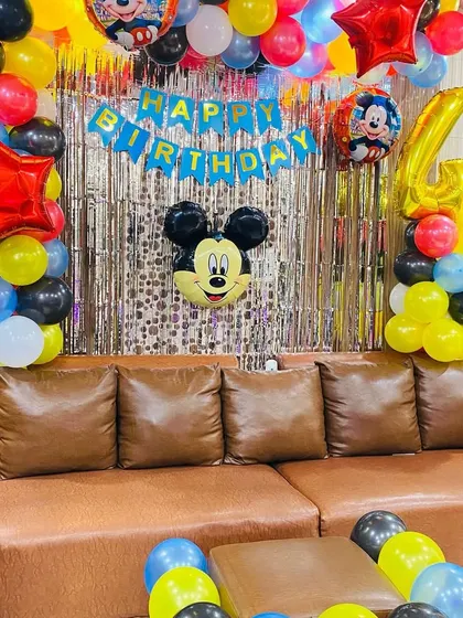 A Mickey Mouse themed 4th birthday party setup in a living room. The decoration includes a balloon arch, character foil balloons, and a silver foil curtain.
