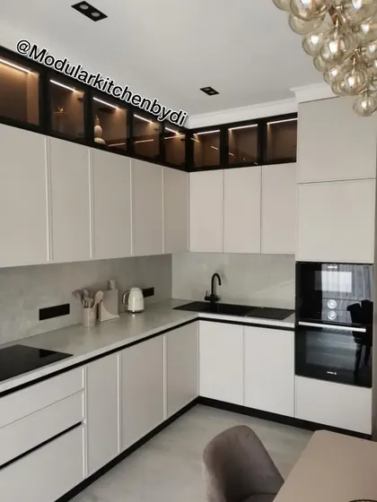 This L-shaped kitchen showcases a beautiful contrast with matte white cabinets, black profile handles, and a dark countertop. The smoked glass upper cabinets with interior lighting add depth and style.