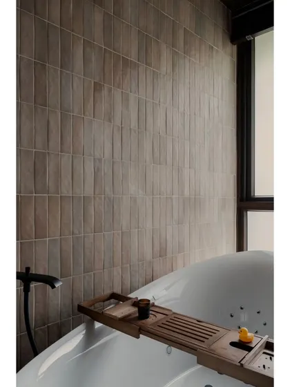 The guest bathroom at Pràna, featuring a freestanding tub and a wall of textured brown tiles. The large window brings in natural light while maintaining privacy.