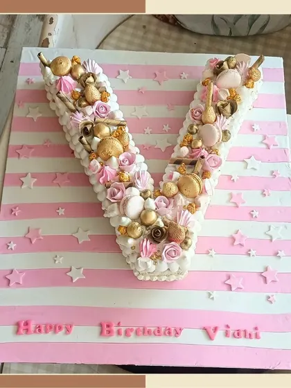 A creative and beautiful monogram cake in the shape of the letter 'V', decorated with macarons, rosettes, and golden pearls.