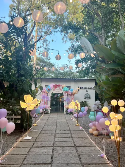 A beautiful, long walkway leading to a party at the 'Backyard' venue. The path is lined with glowing lights and paper lanterns, creating a warm and inviting ambiance for an evening celebration.