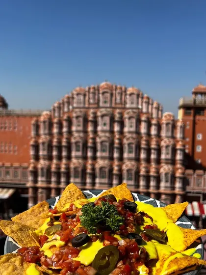 A plate of loaded nachos with cheese sauce, salsa, and olives, framed against the sunny Jaipur sky and Hawa Mahal. This image captures a perfect cafe snack in a beautiful travel setting.