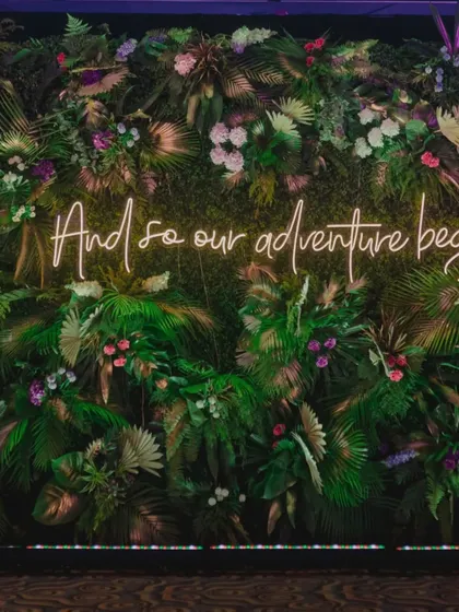 A close-up of the "And so our adventure begins" neon sign against the lush tropical foliage wall.