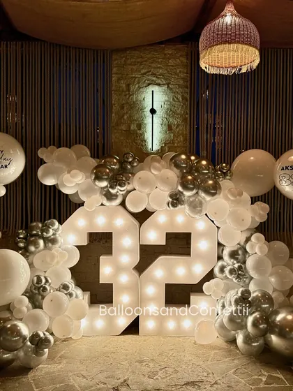 Make your milestone shine. This 32nd birthday setup features large marquee light numbers, framed by a chic white and chrome silver balloon garland, creating a sophisticated and celebratory focal point.