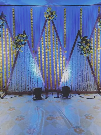 A unique and modern stage design using three large wooden triangles as the backdrop. The triangles are decorated with strings of white and yellow flowers, offering a geometric twist to traditional wedding decor.