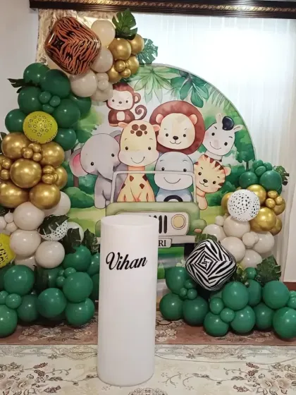 A cute safari-themed decoration for a second birthday. This setup includes a backdrop with cartoon jungle animals, balloon arrangements in green and gold, and personalized name blocks.