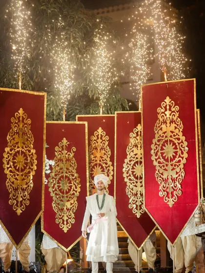 Elegance meets excitement. The groom makes a royal entrance, flanked by attendants holding grand velvet banners and framed by sparkling fireworks.