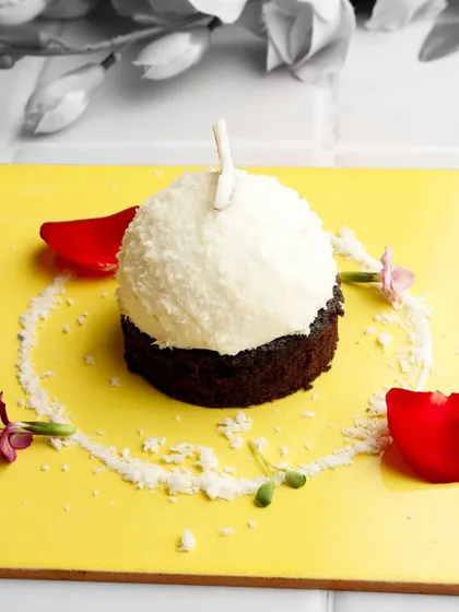 A study in contrast. The pure white of the coconut-dusted dome stands out against the rich, dark chocolate base and the vibrant yellow plate, creating a visually striking dessert.