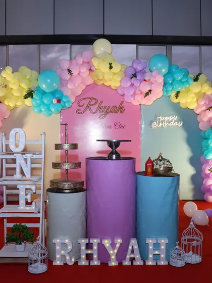 A first birthday party setup featuring a pastel balloon arch, cylindrical plinths for the cake, and large light-up marquee letters spelling out the name 'RHYAH' and a 'ONE' display shelf.