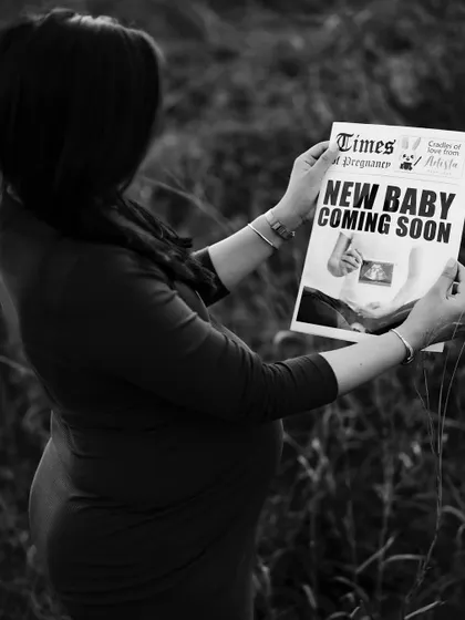 A black and white photo of the mom-to-be reading the "New Baby Coming Soon" newspaper in a field of tall grass, creating a moody and timeless feel.