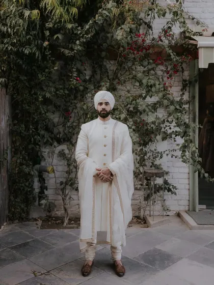 A portrait of a groom in a classic white sherwani, standing against the rustic white brick wall and ivy, exuding timeless elegance.