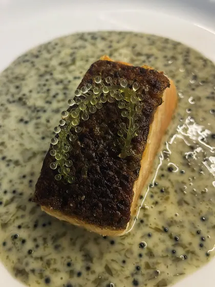 A close-up of pan-seared salmon with perfectly crispy skin. It's served on a bed of creamy sauce and garnished with delicate finger lime caviar.
