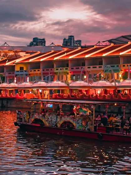 The colorful shophouses of Clarke Quay in Singapore are aglow with lights at dusk, as a river cruise boat passes by on the water.