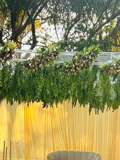 The top border of an outdoor stage, featuring hanging green foliage and floral arrangements against a backdrop of yellow drapes and lush trees.