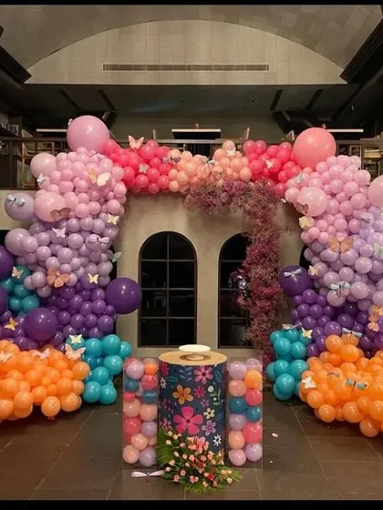 A butterfly garden theme brought to life with a multi-colored balloon arch in shades of orange, purple, pink, and teal. The floral-patterned pedestal for the cake and cascading artificial flowers add to the enchanted forest feel.