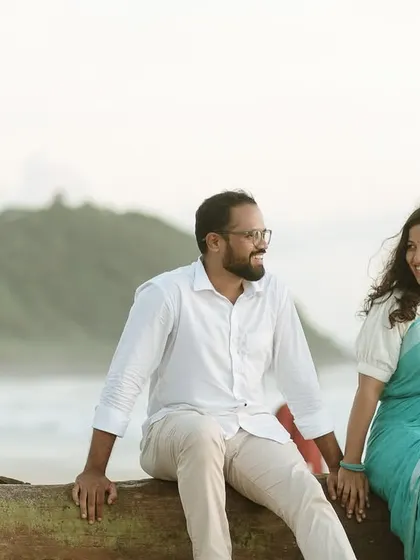 The couple sitting on a rustic log by the sea, sharing a laugh against the beautiful backdrop of the coastline and hills.