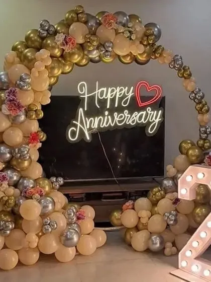 A 25th-anniversary setup with a balloon arch in beige, gold, and silver, framing a 'Happy Anniversary' neon sign.