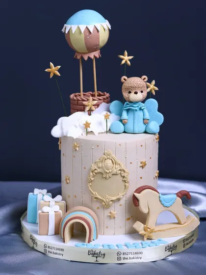 A full view of the whimsical first birthday cake with a teddy bear, hot air balloon, and rocking horse.