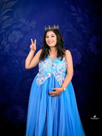 A fun and celebratory solo shot. The mom-to-be, wearing a crown and a beautiful blue gown, flashes a peace sign, sharing her joy and excitement.