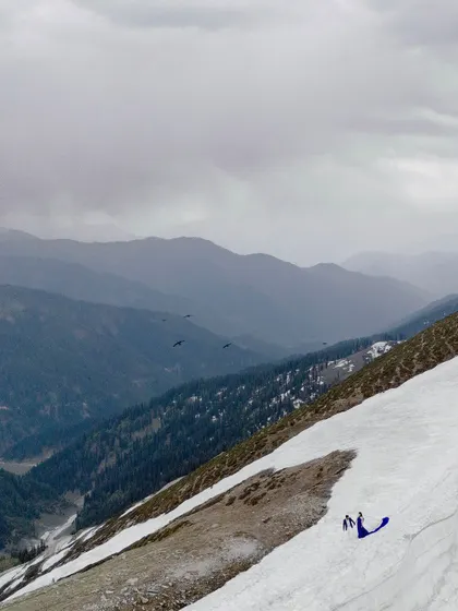 A dramatic wide shot showing the tiny figures of the couple against the vast, snow-covered mountains, highlighting the scale and grandeur of nature.