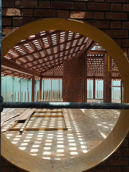 A view through a circular opening in a brick wall during construction. This shot highlights the beautiful play of light and shadow created by a latticed roof, turning a simple material into a dynamic architectural element.