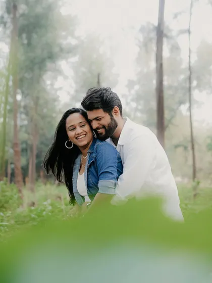 A couple shares a hug in a sun-dappled forest, framed by green leaves in the foreground. This composition creates a sense of intimacy and being lost in their own world.