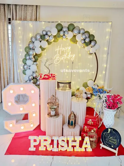 A sophisticated birthday setup with an olive green, white, and silver balloon arch. The large marquee number '9' and plinth arrangement complete the look.