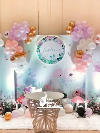 A dreamy photo corner for a butterfly-themed baby welcome. The setup features custom-printed backdrops with floral artwork, butterfly-wing props, and soft balloon arrangements in pink, white, and gold.