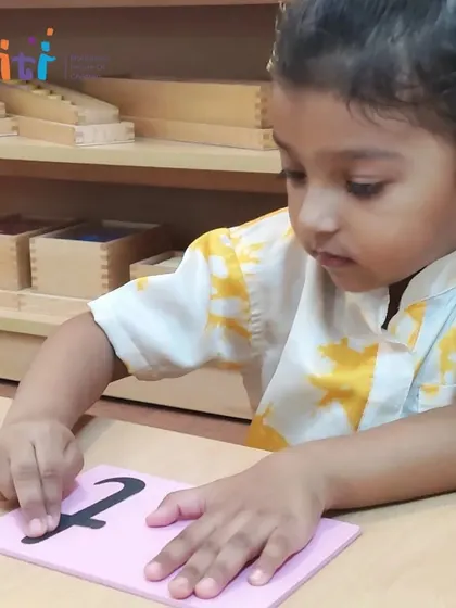 A young girl carefully traces a pink sandpaper letter, representing a vowel sound. This tactile work prepares the hand and mind for writing.