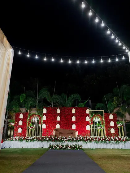A wide shot of our red rose wall stage, set up on a lawn for an outdoor reception. The string lights above and the neat floral border complete the elegant and romantic look.