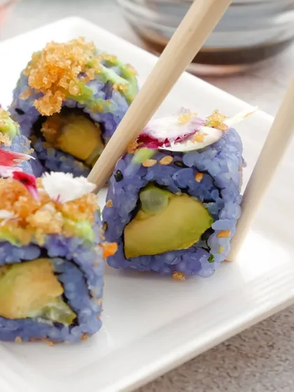 A detailed shot of our unique blue rice sushi. A chopstick picks up a piece, revealing the fresh avocado filling and the delicate edible flower garnish.