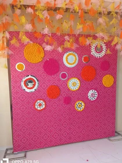 A creative and colorful photo wall for a mehendi or sangeet. The pink patterned backdrop is decorated with circular patterns made from marigold flowers and mirrors.