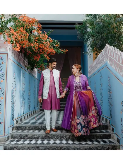 The couple descends a beautifully painted staircase, hand in hand, their colorful attire creating a stunning visual against the blue walls.