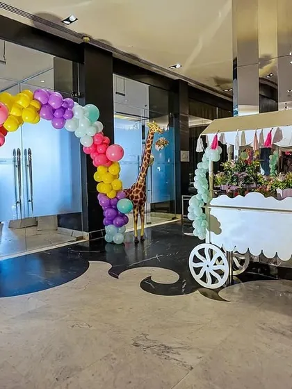 A vibrant entrance to a carnival-themed party. A colorful balloon arch frames the walkway, leading guests past life-sized giraffes and a directional signpost cart, setting a festive tone from the start.