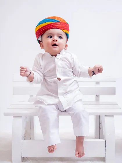 A joyful portrait of a baby dressed in a white kurta and a bright Rajasthani turban. His happy expression and pose on the white bench create a delightful and memorable image.