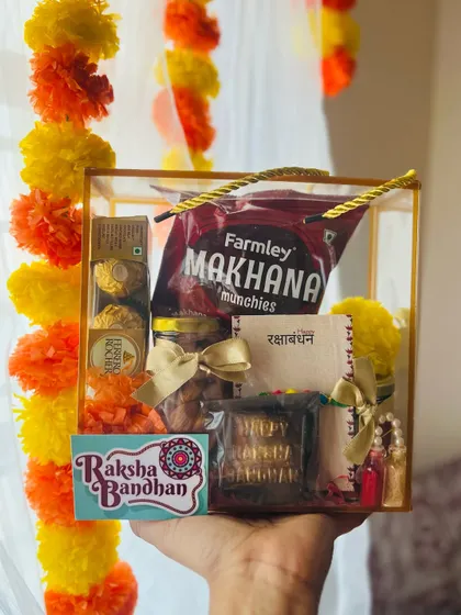 A clear box hamper for Raksha Bandhan, filled with Ferrero Rocher, makhana, and dry fruits.