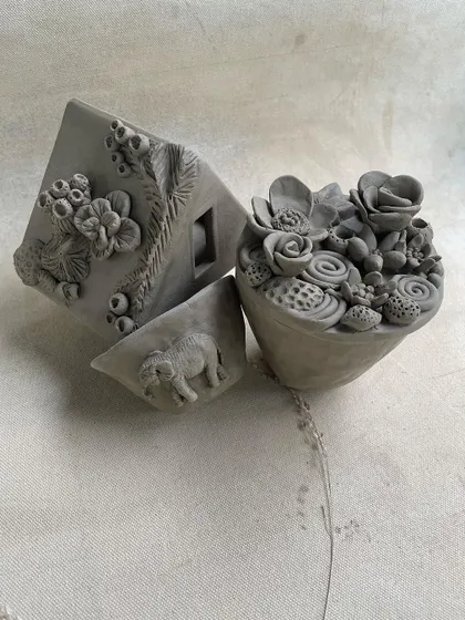 A collection of intricate, unfired hand-built sculptures. This image showcases a house with detailed floral work, a succulent planter, and a piece with an elephant motif.