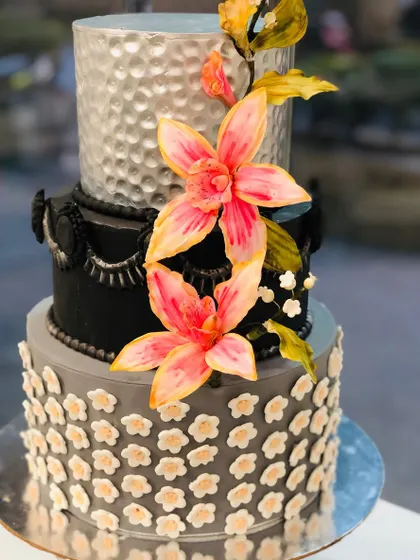 A sophisticated three-tier cake in black, silver, and grey. The design combines different textures on each tier and is finished with striking pink sugar lilies, creating a bold and modern statement.