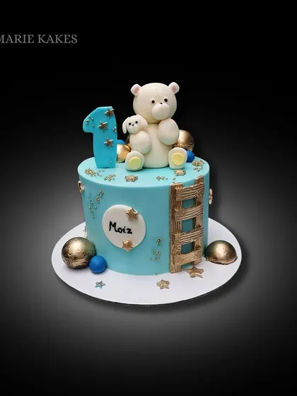 A sweet teddy bear cake for a first birthday. The light blue cake is decorated with gold stars, a little ladder, and an adorable fondant bear topper.