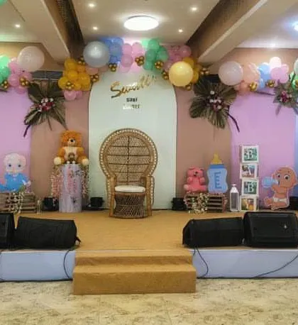 A full stage decoration we did for a large baby shower event. The setup includes a central wicker chair for the mom-to-be, flanked by teddy bear and baby doll props, all set against a pastel pink and blue backdrop.