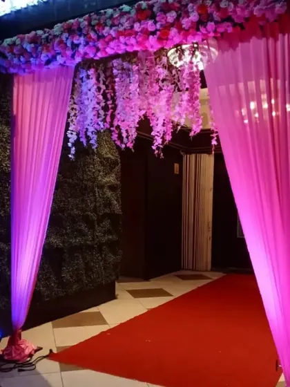 A classic entrance gate decoration for an indoor venue. We used bright pink drapes and an overhead arrangement of artificial flowers with hanging wisteria to create a welcoming and festive entryway.