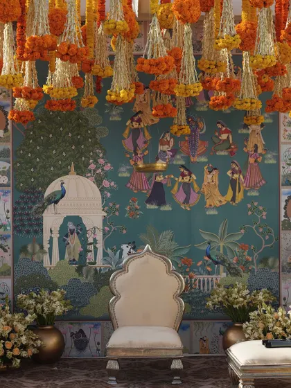 The bride's and groom's chairs are set against a magnificent Pichwai-inspired mural, under a canopy of fresh marigold and tuberose strings.