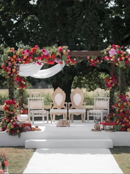 This stunning outdoor mandap combines rustic charm with vibrant tradition. The sturdy wooden frame is adorned with a cascade of bright red, pink, and orange flowers, creating a beautiful and sacred space for a wedding ceremony.