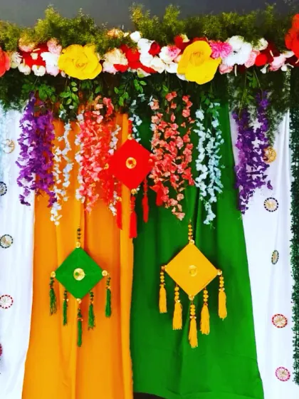 This backdrop was designed specifically for a Teej festival celebration. It features colorful drapes, hanging crafts, and the words 'Teej' and 'Sawan' written out with flowers on green panels.