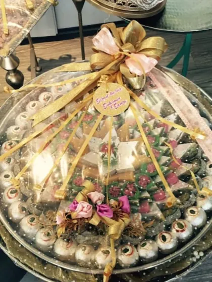 This is an example of a beautifully arranged mithai tray for a wedding. The sweets are artfully placed and wrapped in clear cellophane with a large gold bow, making it a centerpiece of the gift display.