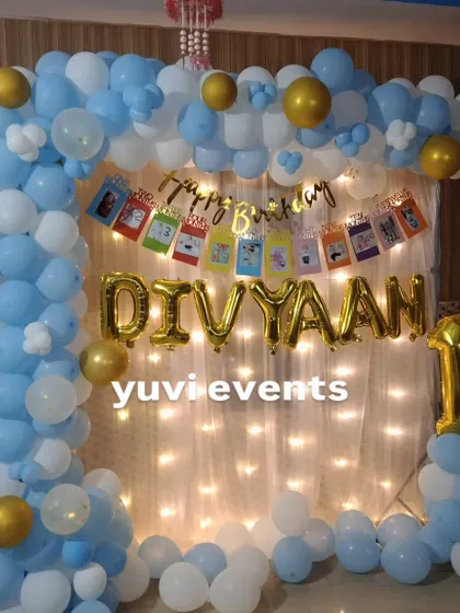 A simple and sweet first birthday decoration at home, with a blue and white balloon frame, fairy lights, and a personalized photo banner.